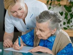 Une professionnelle de santé souriante aide une femme âgée à écrire sur une feuille posée sur une table. La personne âgée tient un stylo pendant que l’aide-soignante se penche à ses côtés pour l’accompagner.
