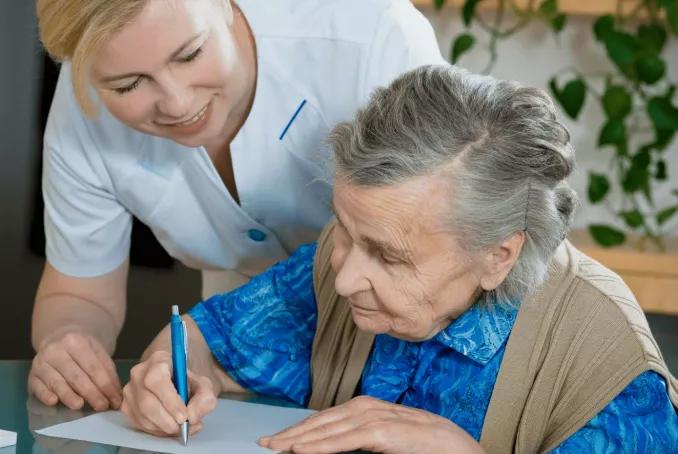 Une professionnelle de santé souriante aide une femme âgée à écrire sur une feuille posée sur une table. La personne âgée tient un stylo pendant que l’aide-soignante se penche à ses côtés pour l’accompagner.